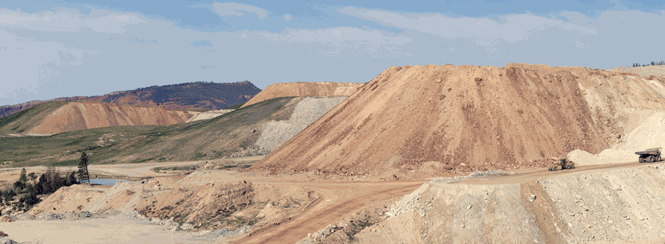Vue d'une mine à ciel ouvert avec de grands monticules de terre beige excavée, une route de terre et un camion minier, sous un ciel bleu parsemé de nuages.