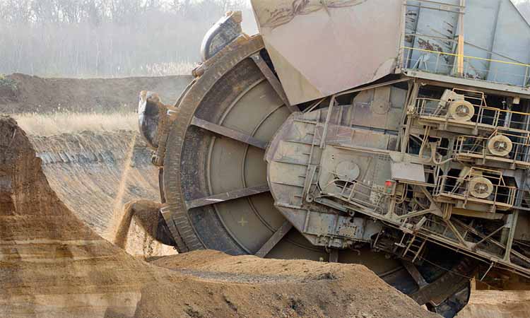 Vue rapprochée d'une grande excavatrice à roue-godets creusant des couches de terre brune et grise, dans une atmosphère poussiéreuse